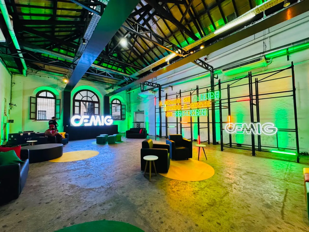 Ambiente em galpão com letreiros em neon, sofás, estrutura metálica e iluminação verde e amarela