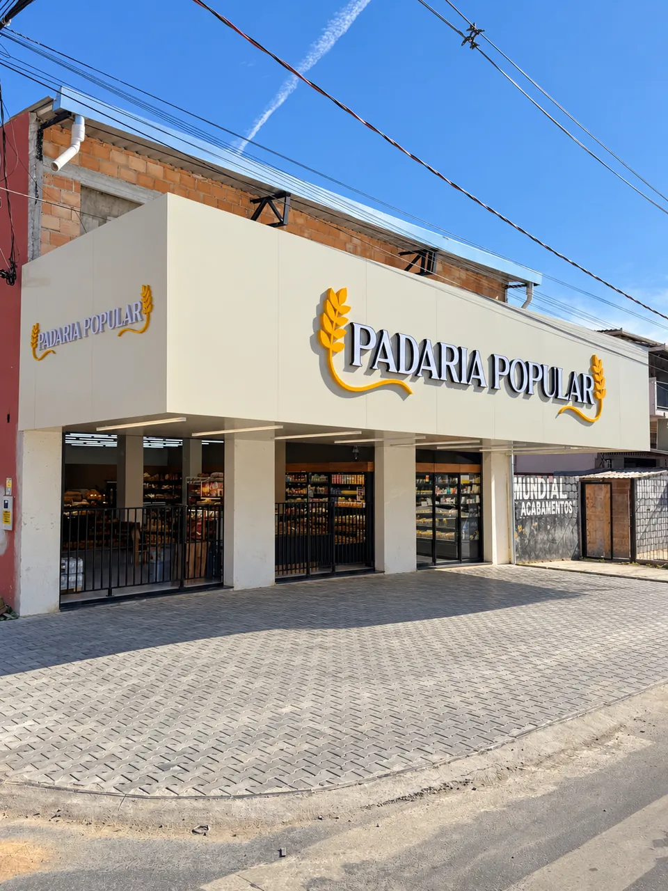 Fachada de padaria em esquina, com letreiro em letras caixa douradas sobre platibanda bege