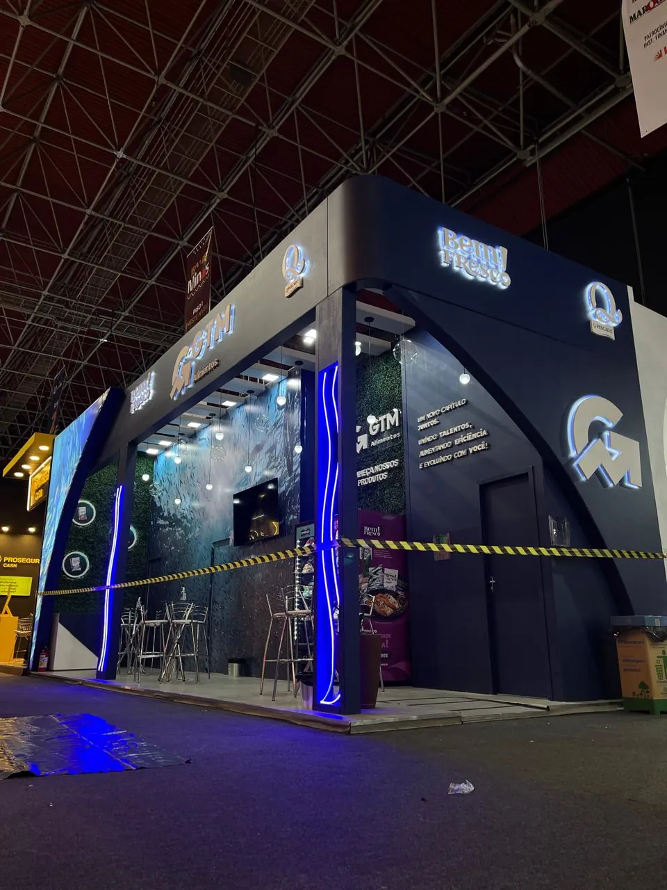 Estande azul-escuro com letreiros iluminados, contornos em LED azul e parede verde, montado em pavilhão de feira
