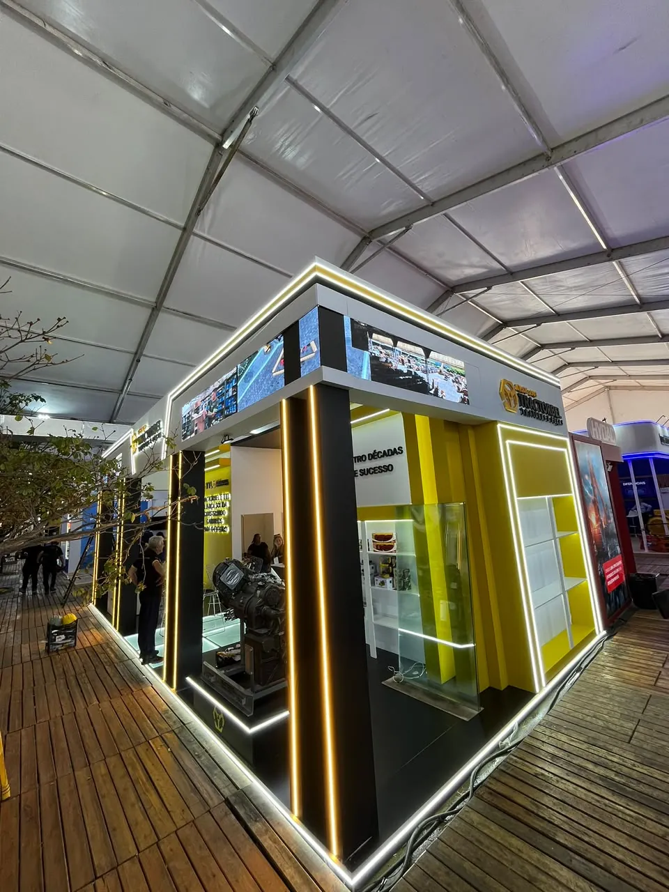 Estande preto e amarelo com contornos em LED e telões no alto, montado sob tenda