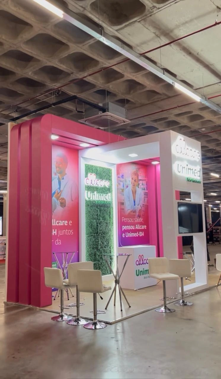 Estande rosa e branco, com poltronas altas e painéis de parceria entre marcas, em pavilhão de feira